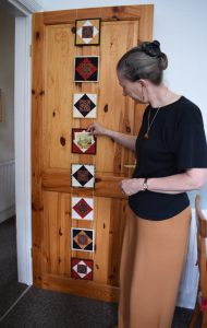 Carolyn standing next to door with red, black and cream square units of patchwork holding cards