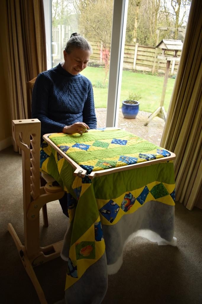 Carolyn Gibbs sitting near window quilting using floor standing hoop.