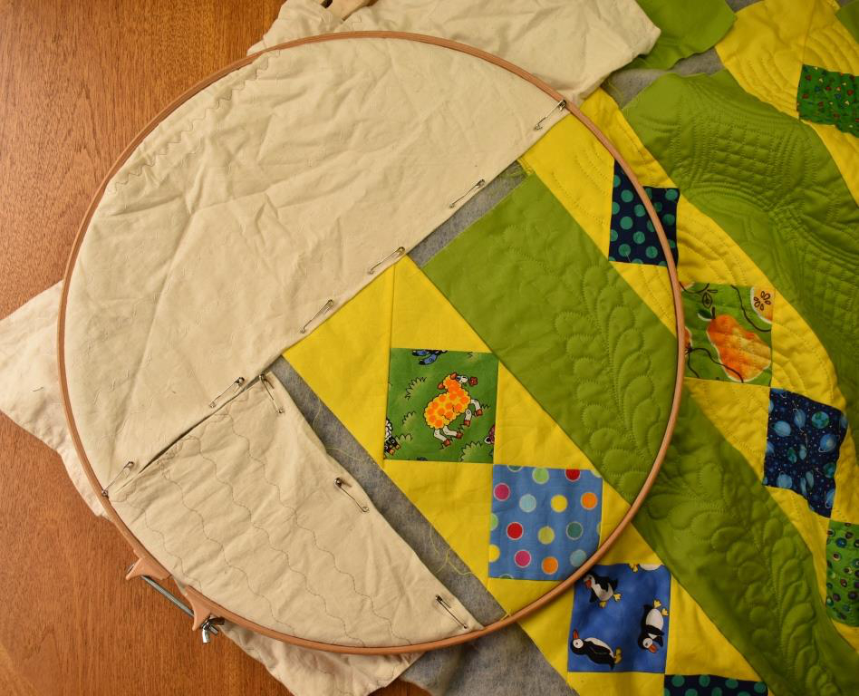 Wide calico strips are pinned onto the edge of the quilting, so that all of the remaining area to be stitched is in the middle of the hoop.